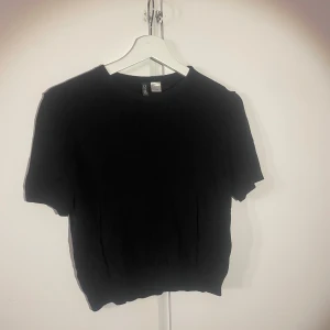 Svart stickad tröja från H&M Divided - En svart, enkel stickad tröja från H&M Divided. Tröjan har rund halsringning, korta ärmar och ribbade kanter. Perfekt till lager-på-lager och funkar året runt. Snygg och basic modell som passar till allt.