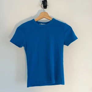 Blå ribbad t-shirt från Zara - Snygg blå t-shirt från Zara i ribbad bomull. Modellen är figurnära med korta ärmar och rund halsringning. Perfekt att matcha med jeans eller kjol för en clean och fräsch look.