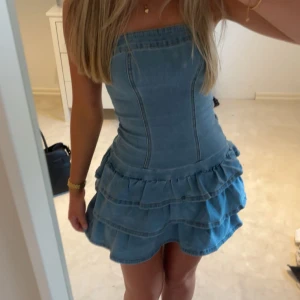 Blå jeansklänning med volanger - Supersöt axelbandslös jeansklänning i blått med figurnära passform upptill och volanger i flera lager på kjolen. Köpt på Plick men är aldrig använd. Stl XS.