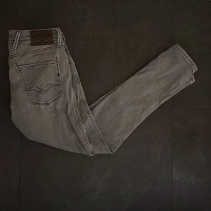Replay Anbass grå jeans, W30 L32 - Snygga Replay Anbass jeans i en ljusgrå färg med klassisk femficksdesign och svarta detaljer på , tillverkade i mjukt bomullsmaterial med lite stretch för extra komfort. Nypris 1800kr