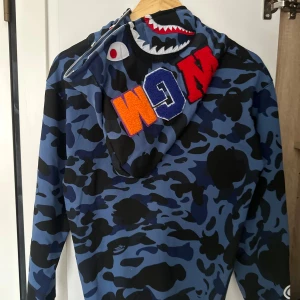 Blå camo hoodie från Bape - Säljer en blå camo hoodie från Bape med ikonisk hajdesign på huvan och broderade bokstäver. Tröjan har hel dragkedja, känguruficka och coola detaljer som tiger- och hajmotiv. Perfekt för dig som vill sticka ut med streetwear.