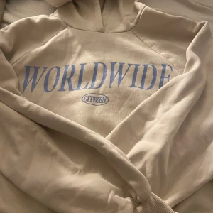 Beige hoodie från Gina Tricot - Säljer en beige hoodie från Gina Tricot med trycket 'WORLDWIDE CITIZEN' i ljusblått på bröstet. Tröjan har huva och långa ärmar, tillverkad i mjuk bomullsmix som är perfekt för chill dagar. Oversized fit för en avslappnad vibe.