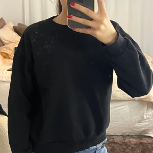 Svart sweatshirt med stjärndetaljer - Svart sweatshirt med rund hals och ribbade muddar. På axeln finns snygga stjärnor i små svarta paljetter som ger en cool touch. Passformen är normal och materialet känns mjukt och skönt, perfekt till jeans. Enkel men ändå med en twist!