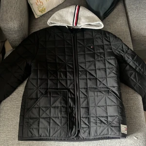 Svart  jacka Tommy Hilfiger - Quiltad svart jacka från Tommy Hilfiger med grå huva och röd-vit-blå rand. Jackan har dragkedja framtill, två fickor och diskret logga på bröstet. Snyggt rutmönster över hela jackan och detaljerad etikett vid nederkanten.