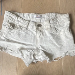Vita jeansshorts från Gina Tricot - Snygga vita jeansshorts från Gina Tricot med fransig kant och slitna detaljer. Klassisk femficksmodell med knapp och dragkedja framtill samt bälteshällor. Perfekta för varma dagar och en avslappnad look.