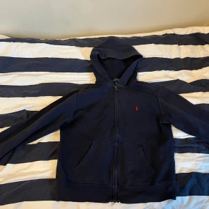 Marinblå hoodie från Polo Ralph Lauren - Snygg marinblå hoodie från Polo Ralph Lauren med röd logobroderi på bröstet. Tröjan har huva, dragkedja framtill och två stora fickor. Tillverkad i mjuk bomull och har långa ärmar. Perfekt för chill dagar och enkel att matcha med jeans. Ena fickan är lite sönder men inget farligt. 