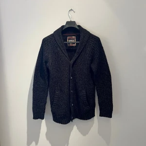 Svart stickad kofta från Stitch & Soul - Mysig svart stickad kofta från Stitch & Soul i storlek M. Koftan har grovstickad struktur, bred krage och knappar framtill. Två fickor på framsidan och långa ärmar gör den perfekt för kyliga dagar. Enkel och stilren design som funkar till många outfits. Hör av er vid fler frågor eller fler bilder 