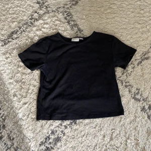 Svart basic t-shirt i bomull - En enkel svart t-shirt med rund hals och korta ärmar. Modellen är rak och har en klassisk passform, perfekt att matcha med jeans eller kjol. Tillverkad i mjuk bomull för en skön känsla hela dagen.