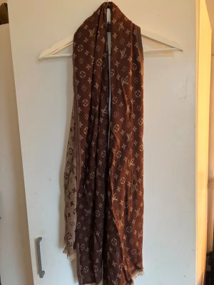 Brun halsduk - Snygg halsduk med klassiskt mönster i beige och brunt. Halsduken har en rak och lång siluett, med fransade kanter och loggor över hela plagget. Perfekt för dig som vill sticka ut med en lux touch och ikonisk design.