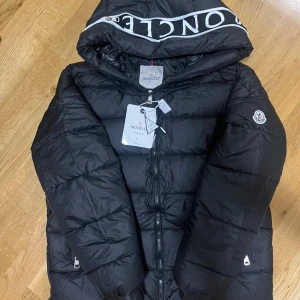 Svart Moncler pufferjacka med huva         L - Svart pufferjacka från Moncler med stor huva och logotyp på ärmen. Jackan har dragkedja framtill, quiltad design och coolt Moncler-tryck på huvan. Insidan har en unik serietidningsetikett. Perfekt för dig som vill ha en snygg och varm jacka med streetkänsla. Fraktar samma dag