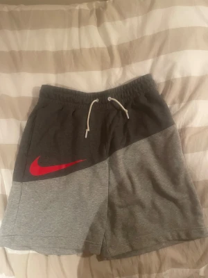 Nike grå shorts med röd swoosh - Snygga grå shorts från Nike med tvådelad färg i mörk- och ljusgrått. De har en stor röd swoosh-logga på benet och vita snören i midjan för justerbar passform. Perfekta för träning eller chill dagar. Mjuk bomullskänsla och avslappnad look.