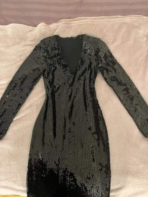 Svart klänning  - Säljer en svart kort klänning med långa ärmar och djup v-ringning. Klänningen är täckt av paljetter som ger en riktigt snygg partylook. Figurnära passform och dragkedja bak. Perfekt för dig som vill sticka ut och glänsa.