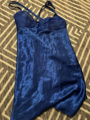 Blå satin linnekläning med spetsdetalj - Säljer en snygg blå linneklänning i satin med tunna axelband och svart spets vid bysten. Toppen har en glansig finish och en figurnära passform som ger en cool och elegant vibe. 