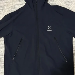 Svart vindjacka från Haglöfs - Svart vindjacka från Haglöfs med huva och två snedställda fickor med dragkedja. Jackan har minimalistisk design och logga på bröstet. Perfekt för utomhusaktiviteter och har hel dragkedja framtill.