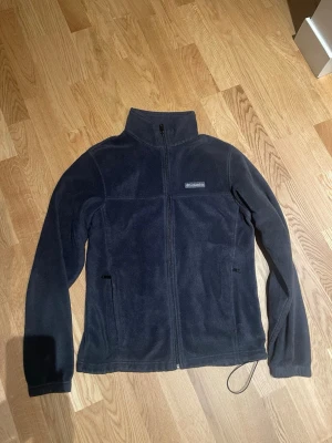 Mörkblå fleecejacka från Columbia stl M män - Mysig mörkblå fleecejacka från Columbia med hög krage och hel dragkedja framtill. Jackan har två sidofickor med dragkedja och justerbar nederkant med snörning. Perfekt för kyliga dagar och outdoor-häng.