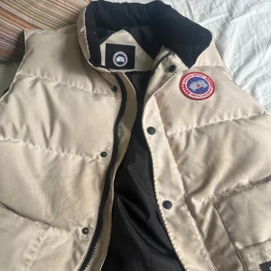 Beige dunväst från Canada Goose - Snygg beige dunväst från Canada Goose med klassisk logga på bröstet. Västen har hög krage, stora framfickor och stängs med både dragkedja och knappar. håller dig varm under kyliga dagar.