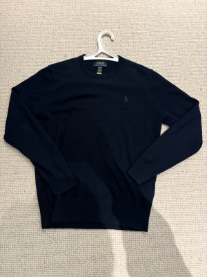 Mörkblå stickad tröja Polo Ralph Lauren - Snygg mörkblå stickad tröja från Polo Ralph Lauren med klassisk rund hals och broderad logga på bröstet. Inga defekter och säljer den eftersom den har blivit för liten. Tröjan har långa ärmar och ribbade muddar vid ärmslut och nederkant. Perfekt för dig som gillar stilrena och tidlösa plagg. 