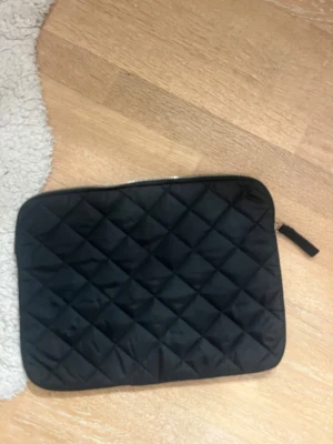 Svart quiltad datorväska - Quiltad svart datorväska från H&M med dragkedja runt om. Väskan har en stilren, rektangulär form och är tillverkad i mjukt syntetmaterial. Perfekt för att bära din laptop på ett snyggt och praktiskt sätt.