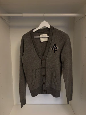 A&F Cardigan - Säljer nu en snygg mörkgrå kofta från Abercrombie & Fitch i storlek L. Koftan har knappar framtill, två fickor och ett broderat AF-märke på bröstet. 