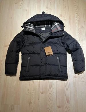 Svart Burberry pufferjacka  - Svart pufferjacka från Burberry med huva och klassiskt rutigt foder i grått, vitt och rött. Jackan har dragkedja, knappar och två fickor med zip framtill. Patch med Burberry London England på ärmen. Perfekt för kalla dagar.