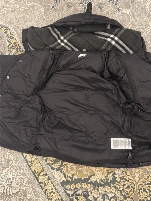Svart Burberry pufferjacka med huva - Säljer en svart pufferjacka från Burberry London England med stor huva och dragkedja framtill. Jackan har quiltad design, tryckknappar och ett rutigt foder i huvan. Perfekt för kalla dagar och har en clean, modern look. Den är också waterproof så att du aldrig blir blött