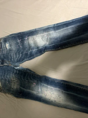 Dsquared2 blå jeans med rosa stänk - Snygga blå jeans från Dsquared2 med slitna detaljer och coola rosa färgstänk. Jeansen har klassisk femficksdesign, tydliga slitningar och en tvättad look som ger en riktigt streetig vibe. Perfekta för dig som vill sticka ut med en unik stil.