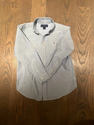 Ljusblå skjorta från Ralph Lauren - Klassisk ljusblå skjorta från Ralph Lauren med button-down krage och broderad logga på bröstet. Skjortan har långa ärmar, knappar framtill och är tillverkad i mjuk bomull. Perfekt för en clean och stilren look.