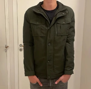 Mörkgrön jacka från John Henric - Snygg mörkgrön jacka från John Henric med quiltad insida, två stora bröstfickor med lock och knappar, samt dragkedja och knappstängning framtill. Jackan har lång ärm och en stilren, rak passform som funkar till många olika looks.
