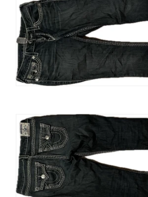 True Religion mörkblåa jeans med kontrastsömmar - Säljer ett par mörkblåa jeans från True Religion med tydliga vita kontrastsömmar och ikoniska fickdetaljer bak. Jeansen har låg midja och raka ben, tillverkade i klassisk denim. Snygga detaljer med knappar och ficklock, perfekt för dig som gillar streetstyle. Bra skicka förutom lite lite slitage där bak, långa i benen! 