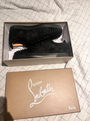 Christian Louboutin svarta sneakers med nitar - Säljer ett par svarta sneakers från Christian Louboutin i mocka med ikoniska röda sulor. Skorna har svarta nitar på tån, rund tå, snörning och coola detaljer. Kommer med originalkartong, dustbag och extra tillbehör. Perfekta för dig som vill sticka ut.