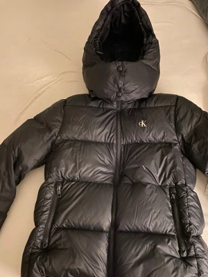 Svart pufferjacka från Calvin Klein - Svart pufferjacka från Calvin Klein Jeans med huva och två dragkedjeförsedda fickor. Jackan har en quiltad design och CK-logga på bröstet. Perfekt för kalla dagar med sin tjocka och fluffiga look. Stängs med dragkedja framtill och har en modern, clean stil.