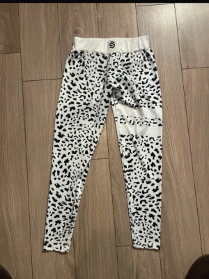 Vita leopardmönstrade leggings - Snygga vita leggings från STRONGER med svart leopardmönster. De har hög midja och är tillverkade i ett stretchigt syntetmaterial som passar perfekt för träning eller chill. Superskön passform och cool design som sticker ut.