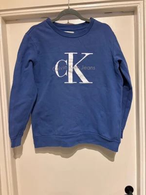 Blå Calvin Klein sweatshirt. - Köpt på Vinter. Passform ej som önskat Skick: Använd men fint. Vissa sprickor i mönster samt ett hål vänster arm, se bild.   Tvättråd: 30°