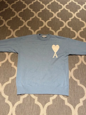 Ljusblå stickad tröja från Ami - Ljusblå stickad tröja från Ami med rund hals och ribbade muddar. På bröstet finns ett stort vitt hjärta med bokstaven A. Tröjan är i mjuk ull och har en clean, stilren look som passar perfekt till jeans eller chinos.