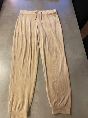 Beige mjukisbyxor med snörning - Snygga beige mjukisbyxor med ribbade muddar vid bensluten och justerbar snörning i midjan. Materialet är mjukt och har en lätt glansig finish, vilket ger en chill och skön vibe. Perfekt för att hänga hemma eller dra ut på stan. Säljer då dom är för stora för mig.