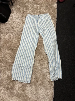 Randiga bomullsbyxor från Lindex - Säljer ett par sköna och luftiga byxor från Lindex i 100% bomull. De är vita med blå och ljusgula ränder, har elastisk midja med snörning och raka ben. Perfekta för chill dagar eller när du vill ha något bekvämt på dig.