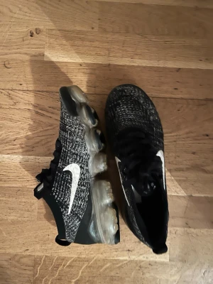 Nike Vapormax Flyknit svart/vit - Nike Vapormax Flyknit sneakers i svart och vit med stickad ovandel och tydlig vit swoosh på sidorna. Skorna har genomskinlig plattformsula med synliga luftkuddar och snörning framtill. Perfekt för dig som gillar sportig och modern stil.