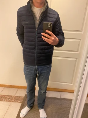 Mörkblå pufferjacka från Timberland - Säljer en mörkblå pufferjacka från Timberland i storlek M. Jackan har quiltad design, dragkedja framtill och stårkrage. Tillverkad av återvunnen polyester och är lätt att packa ner. Perfekt för dig som vill ha en clean och funktionell look.