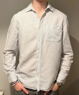 Randig vit skjorta från Zara - Fräsch vit skjorta från Zara med tunna blå ränder och klassisk krage. Skjortan har lång ärm, bröstficka och knappar framtill. Tillverkad i lätt bomull som känns skön mot huden. Perfekt för dig som gillar stilrena och enkla plagg med lite edge. Pris kan möjligen diskuteras.