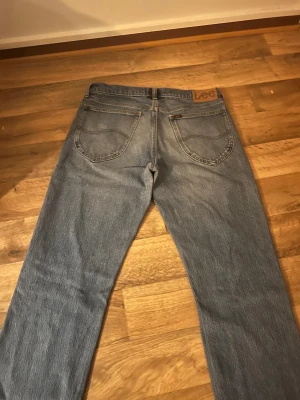 Lee ljusblå raka jeans - Säljer ett par klassiska ljusblå jeans från Lee med raka ben och fem fickor. Jeansen har normal passform och är tillverkade i slitstarkt bomullsjeansmaterial. Snygga detaljer med Lee-logga på bakfickan och metallknapp framtill.