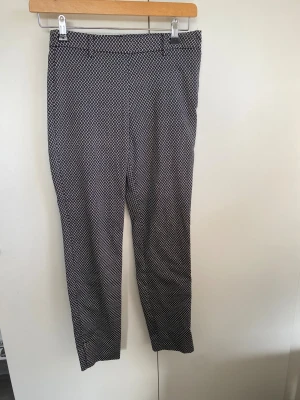 Svarta mönstrade kostymbyxor - Snygga svarta kostymbyxor med vitt geometriskt mönster. Byxorna har rak passform, bälteshällor och diskreta bakfickor. Materialet känns mjukt och har en lätt stretch. Dragkedja i sidan för smidig påklädning. Perfekta för dig som gillar stilrena detaljer.
