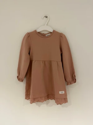 Newbie klänning - 86 Storlek - Beige klänning från Newbie i mjuk bomull, med långa puffärmar och rund hals. Klänningen har fina spetsdetaljer längst ner och knappar i nacken för enkel påklädning. 