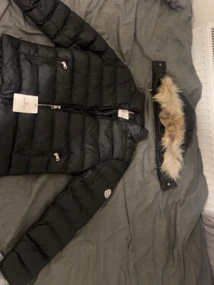 Svart Moncler dunjacka med päls - Svart dunjacka från Moncler med avtagbar huva och snygg pälsdetalj. Jackan har två framfickor med dragkedja, Moncler-logga på ärmen och quiltad design. Perfekt för kalla dagar och har en stilren look som funkar till allt.