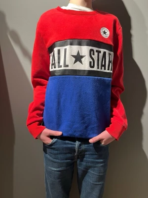 Röd och blå Converse All Star tröja - Snygg röd och blå tröja från Converse med stort 'ALL STAR'-tryck över bröstet och klassisk Converse-logga på vänster sida. Tröjan har lång ärm, rund hals och är gjord i mjuk bomull. Perfekt för dig som gillar streetwear och vill sticka ut med färg.
