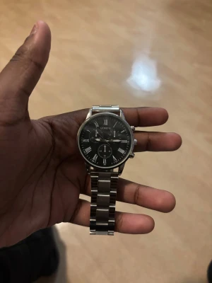 Stilren silverfärgad klocka Geneva - Snygg klocka från Geneva med svart urtavla och romerska siffror i vitt. Armbandet är i blankt, rostfritt stål med klassisk länkdesign. Klockan har tre dekorativa subdials och en rund boett. Perfekt för dig som gillar en clean och tidlös look.