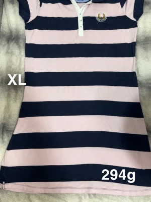 Randig klänning XL från marine classics - Säljer en klänning från marine classics i storlek XL. Klänningen har breda rosa och marinblå ränder, korta ärmar och en klassisk krage med knappar framtill. Den har en broderad logga på bröstet.🌸 Använd en gång. Storlek XL