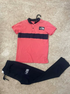 Rosa och marinblå väst från The North Face - Snygg väst från The North Face i rosa med bred marinblå rand över bröstet och marinblå krage. Västen har kort ärm, knappar framtill och logga på vänster sida. Tillverkad i mjuk bomull som känns skön mot huden. Perfekt för dig som gillar sportig och clean stil.