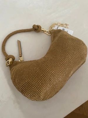 Guldig axelväska från Steve Madden - Säljer en snygg axelväska från Steve Madden i guld med glittriga paljettdetaljer och chunky kedja. Väskan har en cool böjd form, dragkedja och ett dekorativt SM-hänge. Materialet är syntet och hela looken är riktigt statement.