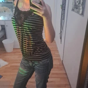 Randig svart linne med spetsdetalj - Säljer ett svart linne med tunna axelband och horisontella gråa ränder. Toppen har en snygg spetsdetalj vid urringningen som ger en extra touch. Passformen är normal och toppen är ärmlös, perfekt för varmare dagar eller lager på lager.