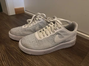 Nike Air Force 1 Flyknit 2 White Pure Platinum - Säljer ett par fräscha Nike Air Force 1 Flyknit sneakers i helvitt. Skorna har klassisk låg siluett, stickad ovandel i flyknit-material och vita snören. Perfekta för dig som gillar stilrena och lätta sneakers med ikonisk design.